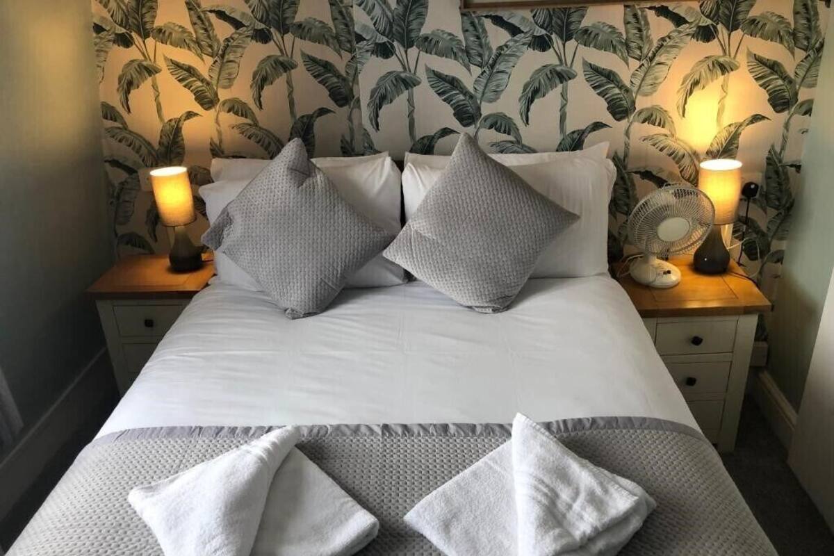 A neatly made bed with two pillows, a bedside lamp, and a fan on a wooden nightstand.