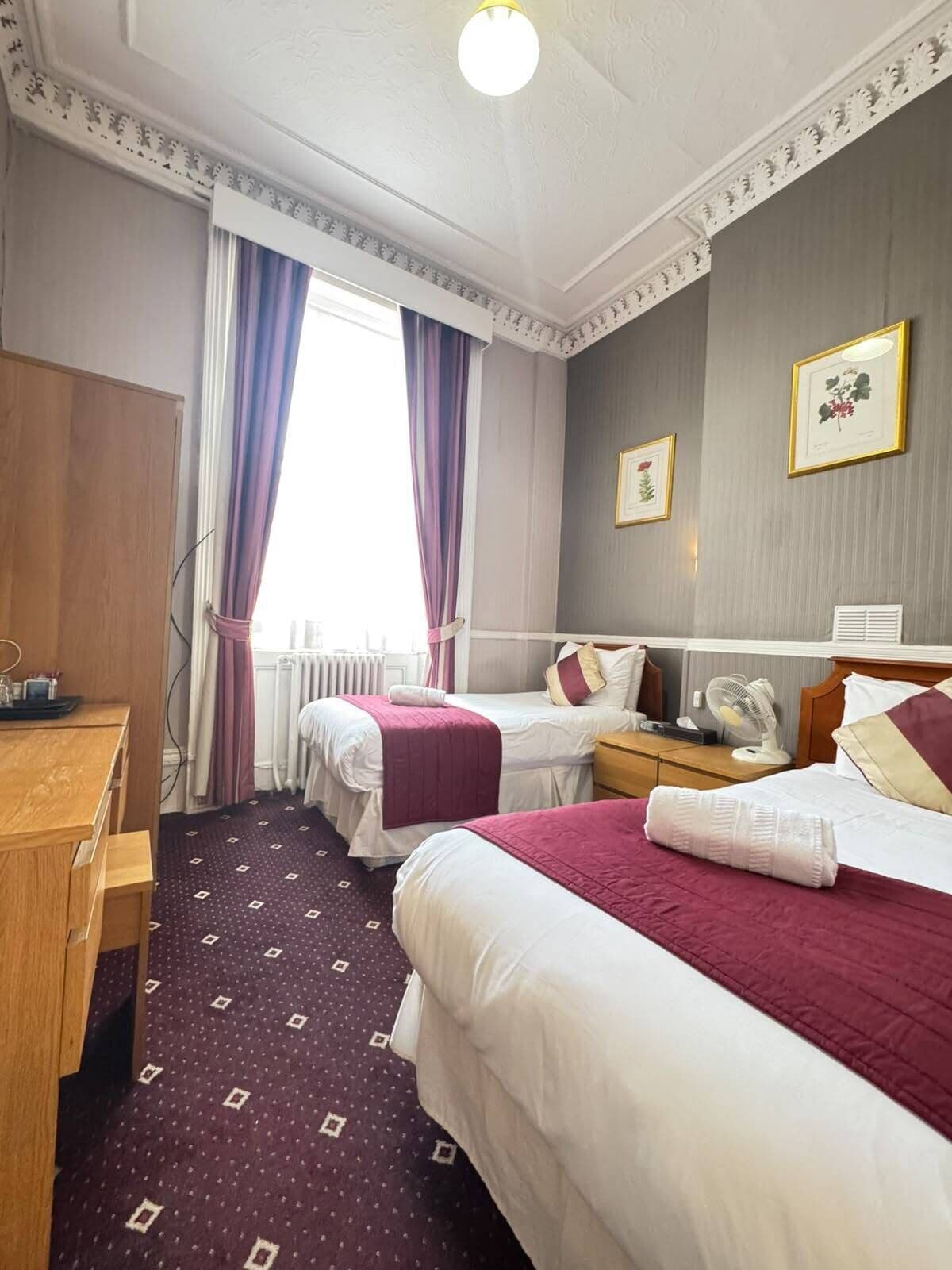 A hotel room with two beds, a desk, a chair, a radiator, and a window with curtains.