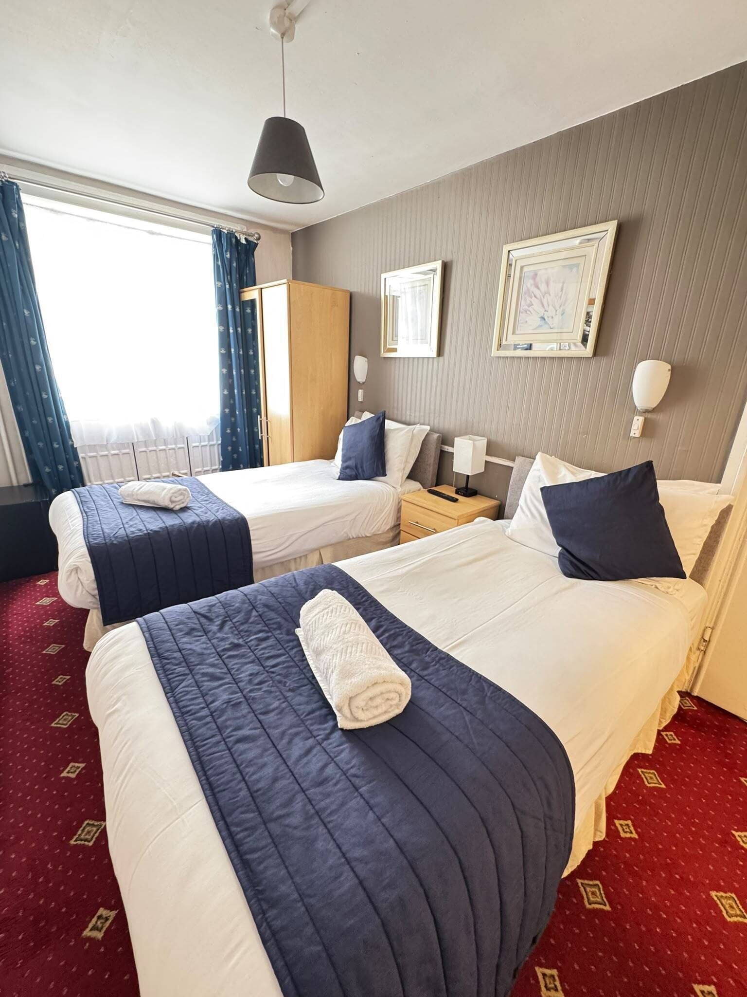 A hotel room with two beds, a wardrobe, a bedside table with a lamp, and a window with curtains.