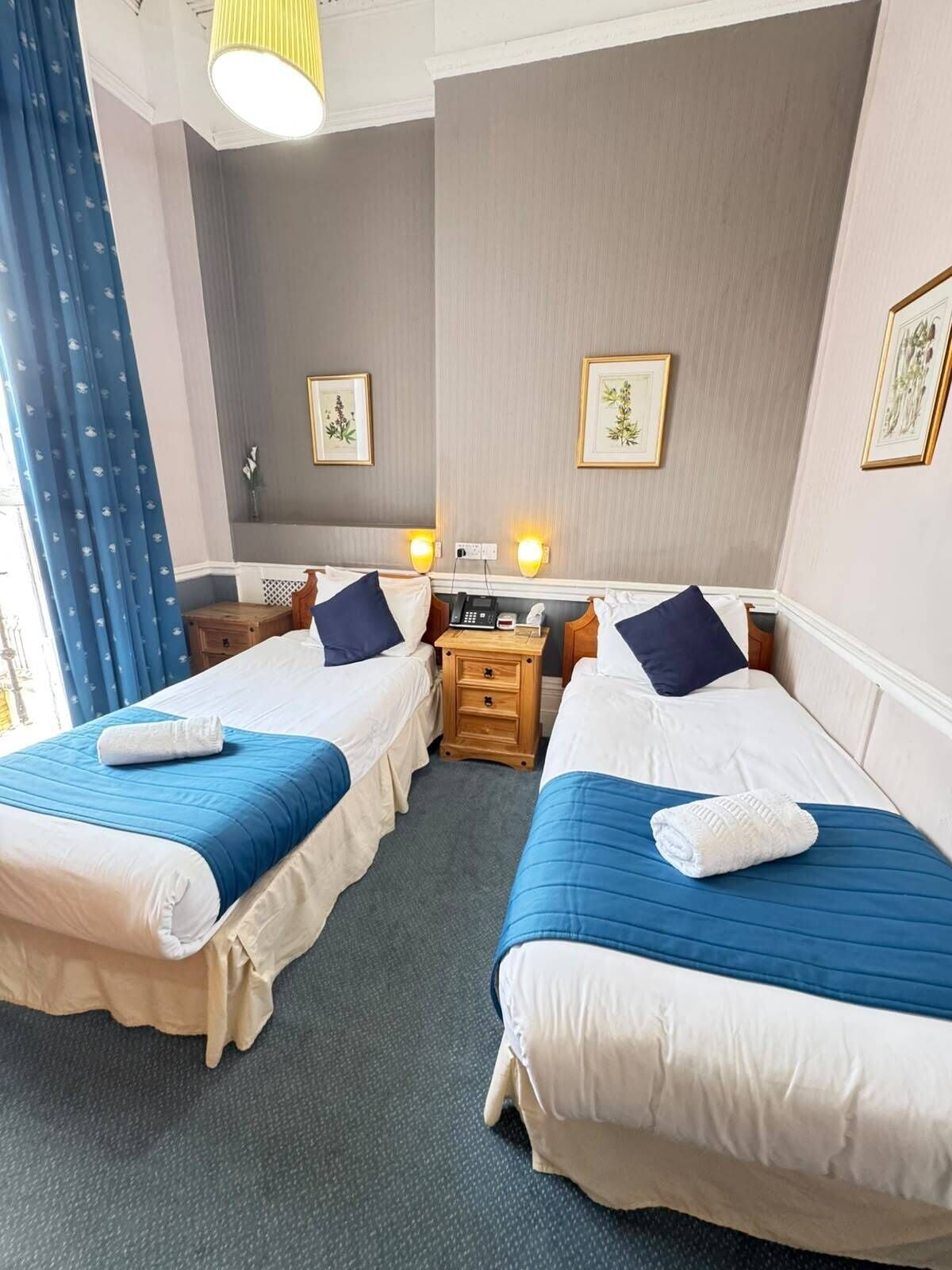 A hotel room with two beds, a bedside table, a lamp, and framed artwork on the wall.