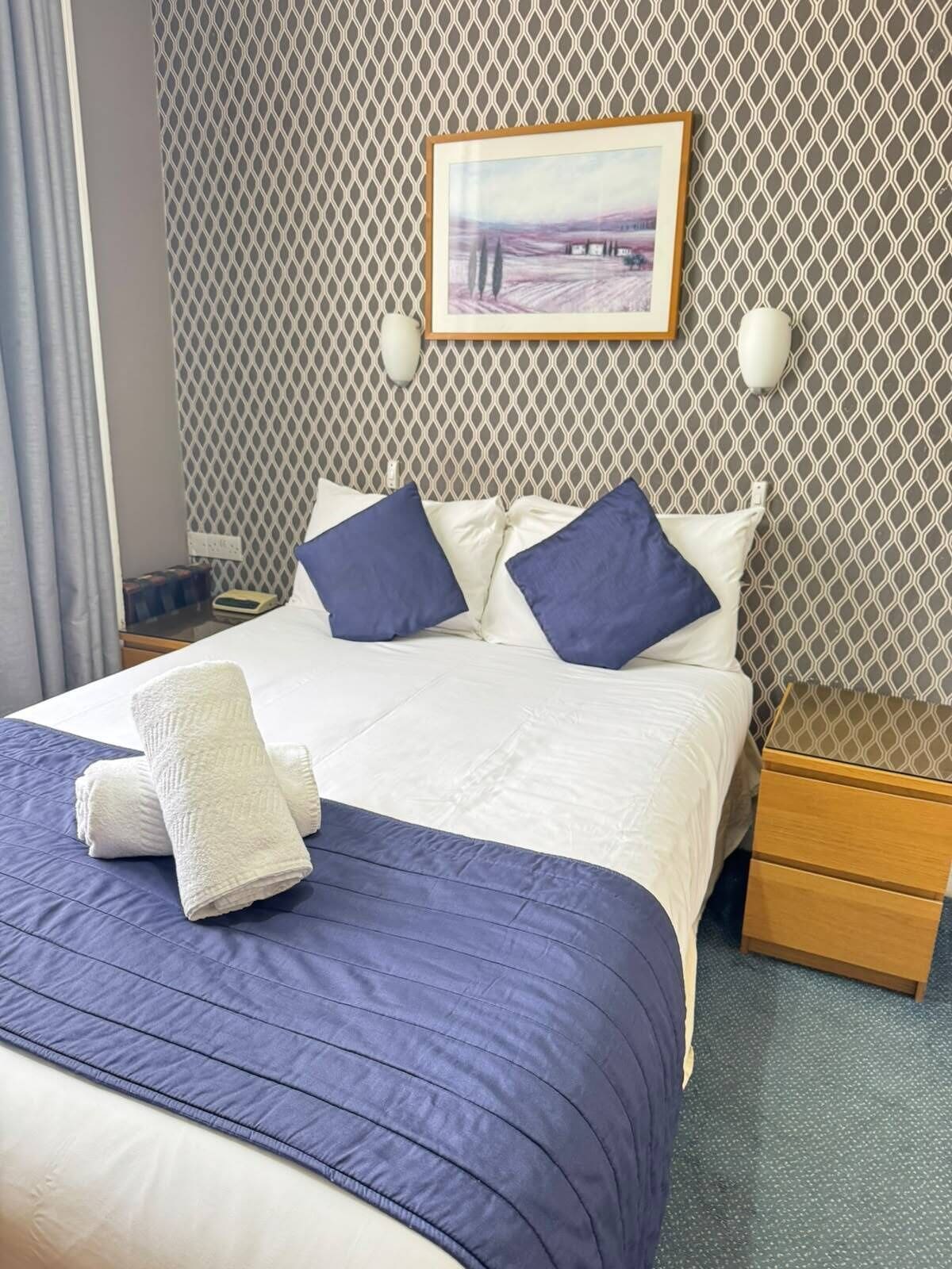 A neatly made bed with a blue and white duvet, two pillows, and a folded white towel.