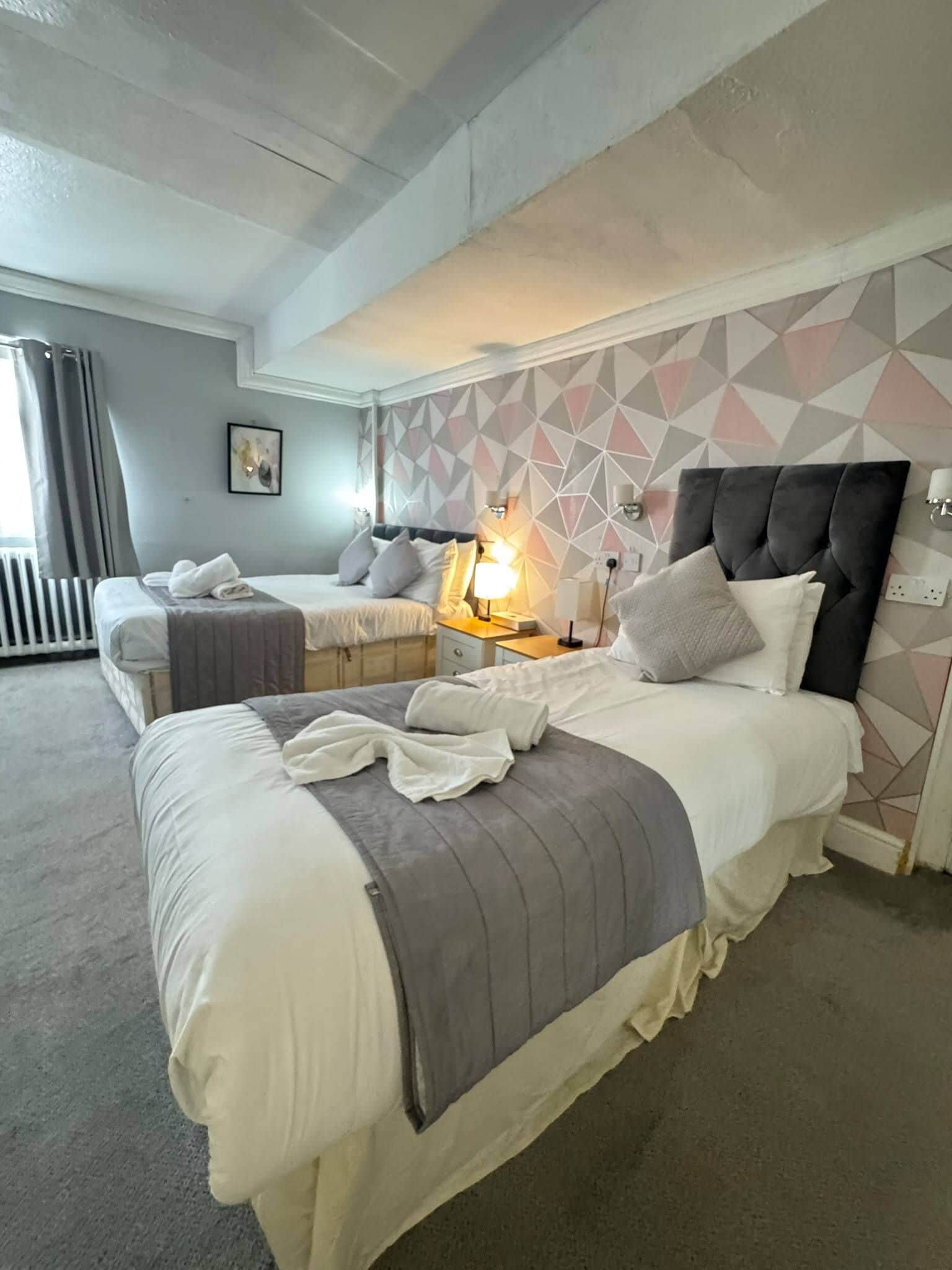 Habitación de hotel con dos camas, un radiador y una lámpara de noche, además de un papel tapiz con diseño geométrico.