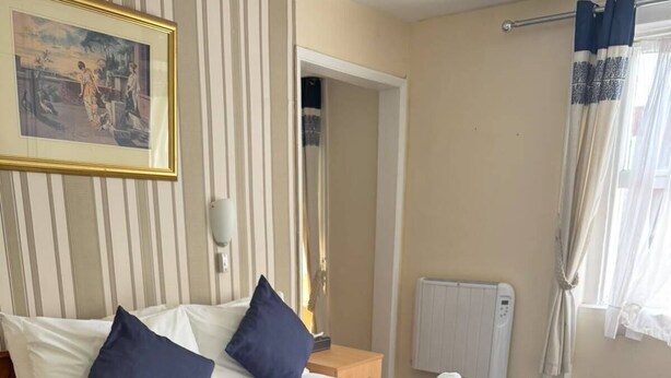 A hotel room with a large bed, two pillows, a folded towel, a radiator, a painting on the wall, and a window with curtains. - Parkwood at Marble Arch (London)