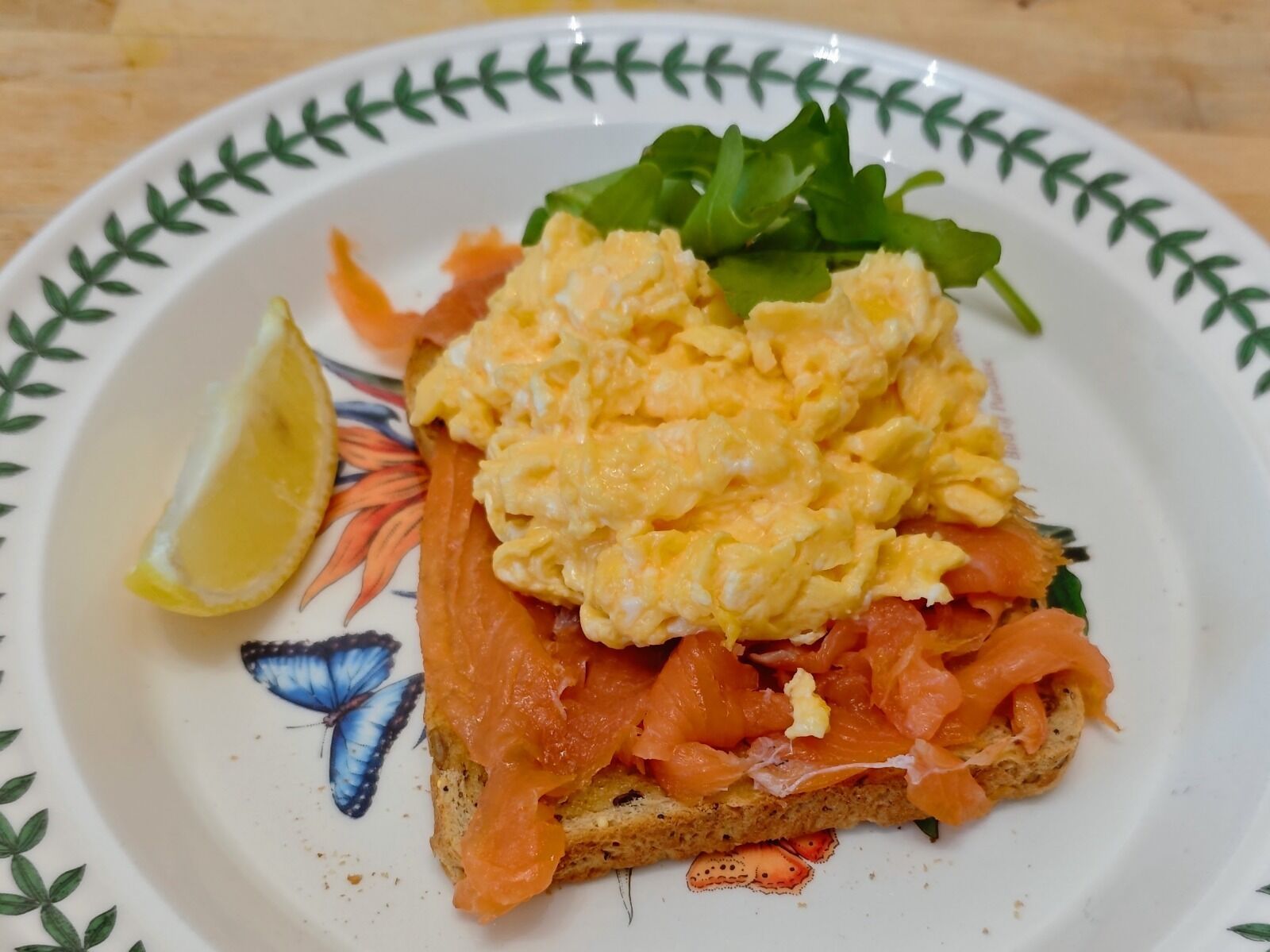 Sándwich con huevos revueltos y salmón ahumado sobre una rebanada de pan, acompañado de una rodaja de limón y algunas verduras en un plato decorativo.