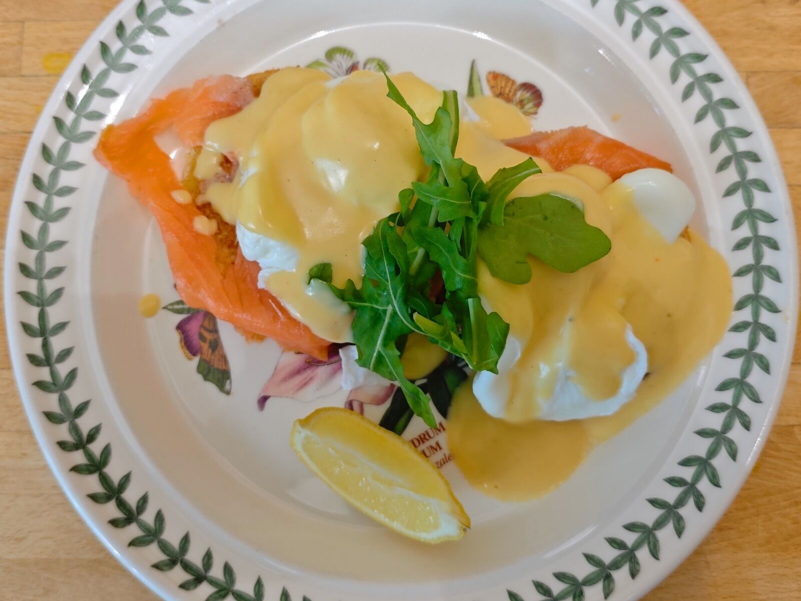 Un plato con salmón ahumado, salsa holandesa, huevos pochados y rúcula sobre un plato decorativo.