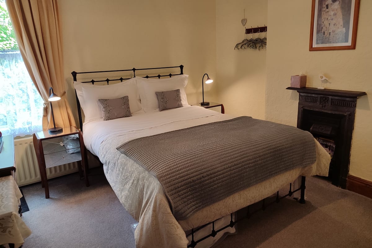 Une chambre à coucher bien rangée, avec un grand lit, des tables de chevet, une lampe, une cheminée et un tableau encadré au mur.