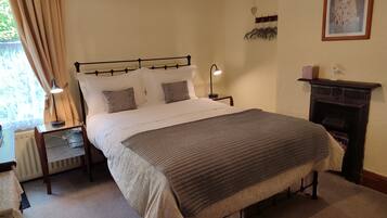 A neatly arranged bedroom with a large bed, bedside tables, a lamp, a fireplace, and a framed picture on the wall.
