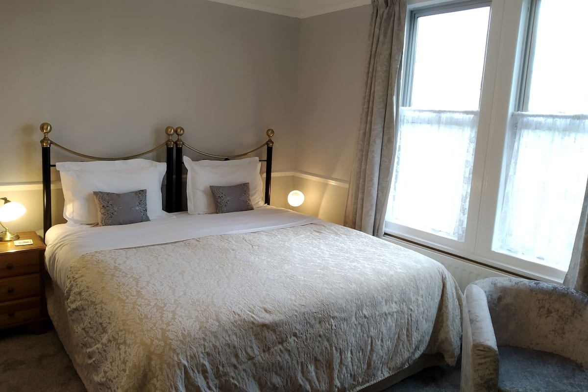 Un lit bien fait, recouvert d’une couvre-lit à motifs, une table de chevet avec une lampe et une fenêtre avec des rideaux.