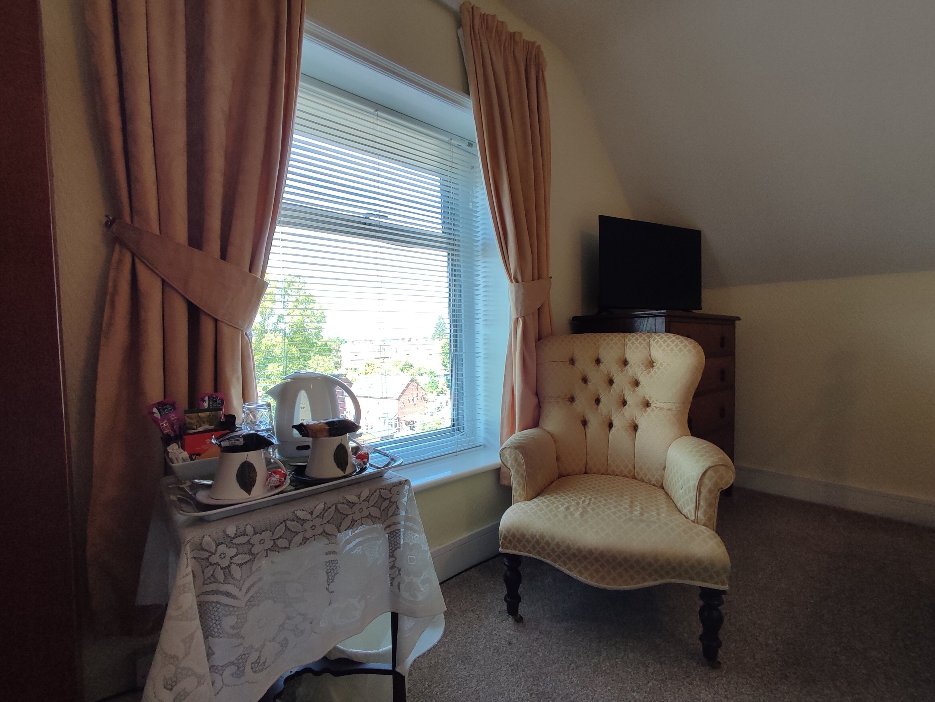 Una habitación con ventana, una mesa con mantel de encaje, una silla y un televisor sobre un soporte.