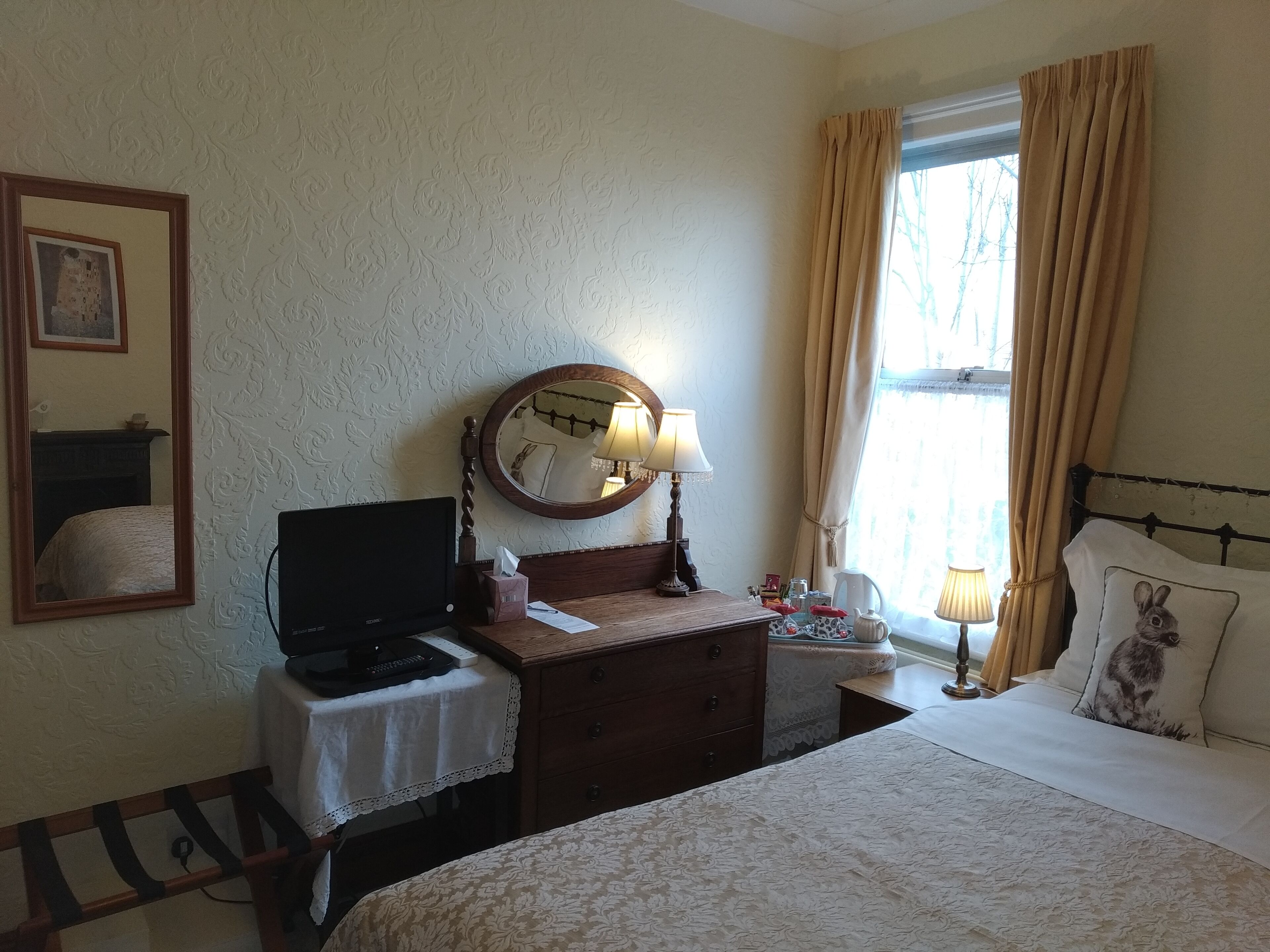 A hotel room with a bed, a desk with a computer, a mirror, a window with curtains, and a bedside table with a lamp.