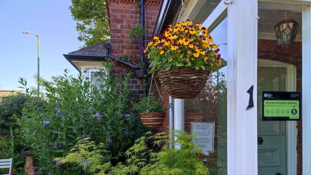Un bâtiment à la façade en briques, une jardinière suspendue remplie de fleurs jaunes et violettes, et une porte sur laquelle est apposée une affiche indiquant le niveau d’hygiène alimentaire.