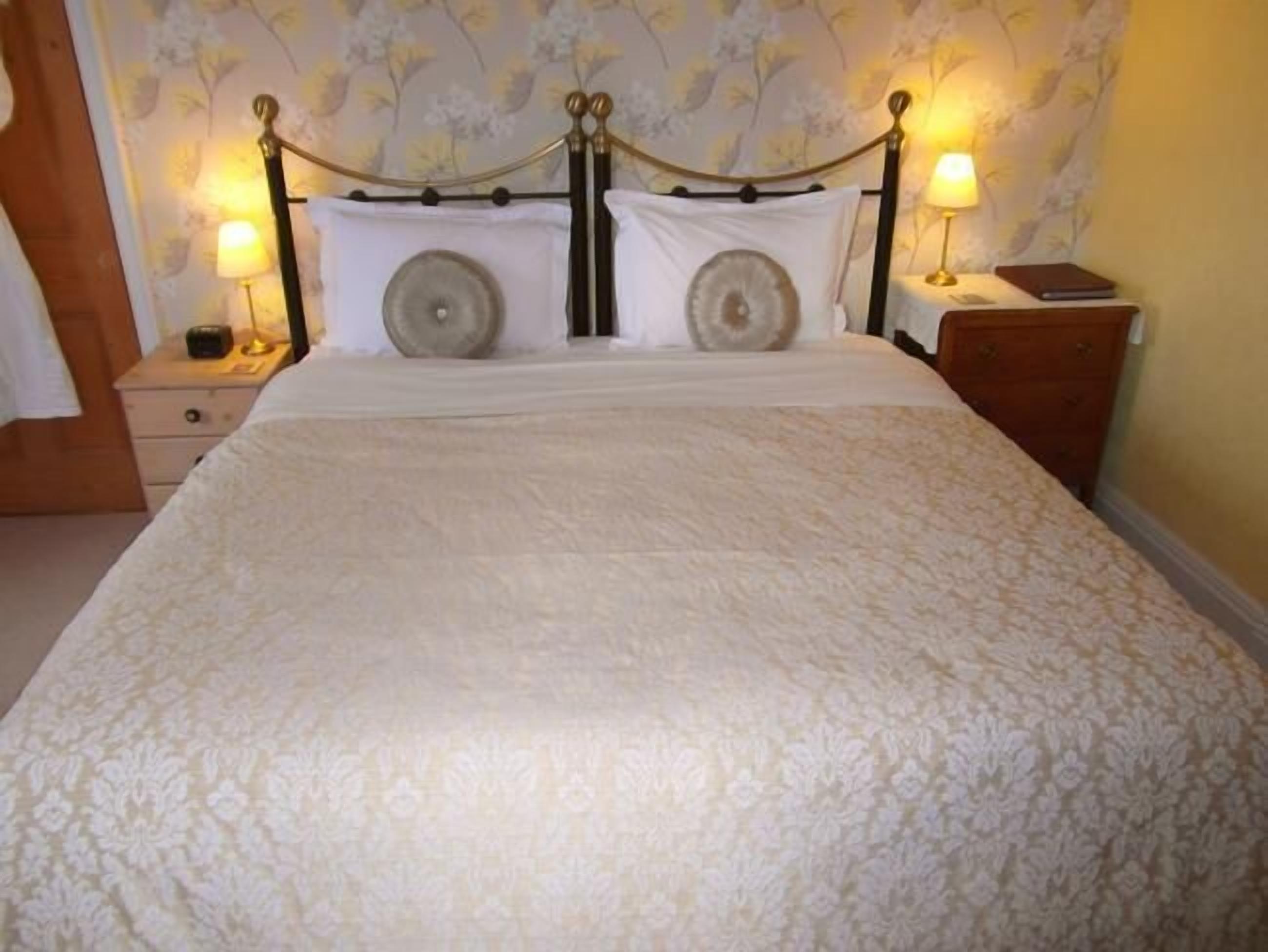 Una cama bien hecha con un cabecero floral, dos lámparas de noche y una mesita de noche con un libro y una lámpara.