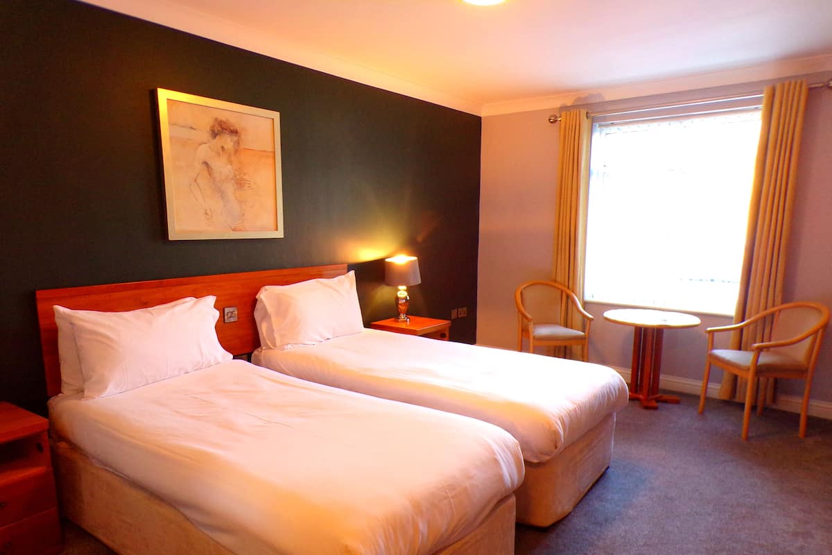A hotel room with two beds, a small table, two chairs, a lamp, and a painting on the wall.
