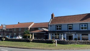 Un pub llamado 'The Fox & Goose' con fachada de piedra, techo de tejas y mesas exteriores con sombrillas.