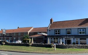 Un pub llamado 'The Fox & Goose' con fachada de piedra, techo de tejas y mesas exteriores con sombrillas.
