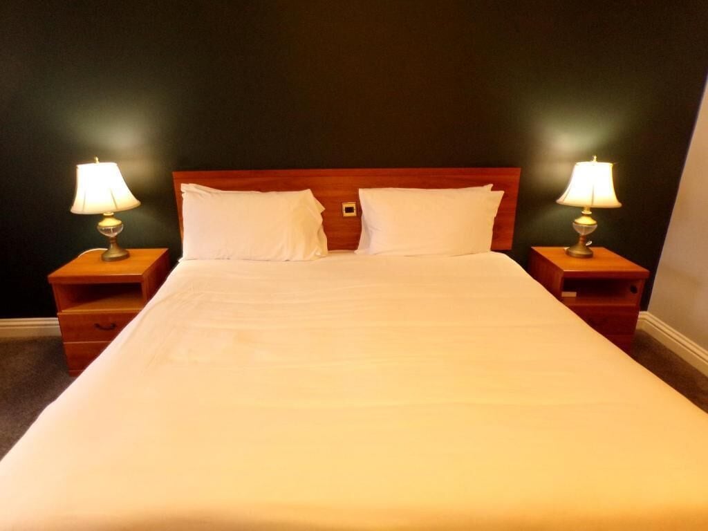 Een netjes opgemaakt bed met witte lakens, twee nachtkastjes met lampen en een donkere muur.