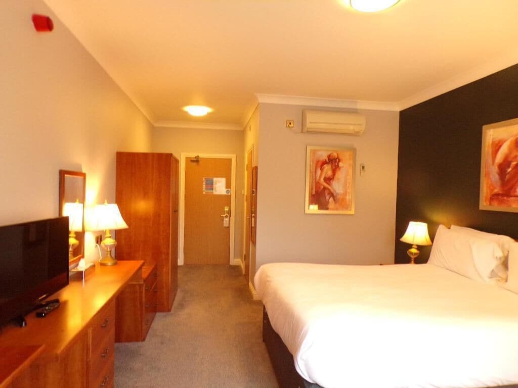 A hotel room with a bed, a TV, a desk with a lamp, a wardrobe, and a door leading to another room.