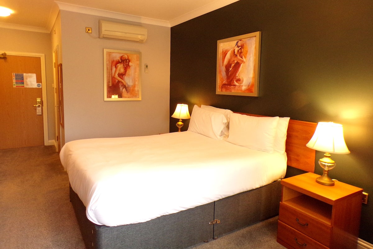 A hotel room with a bed, two bedside lamps, a wooden nightstand, a door, and two paintings on the wall.