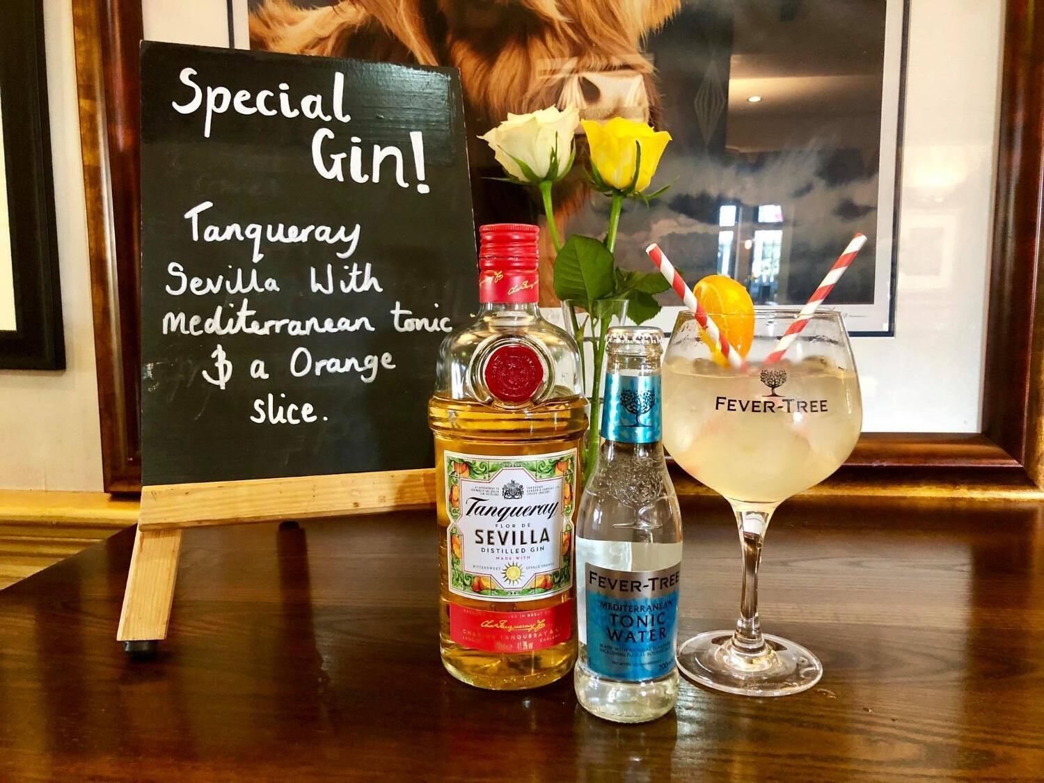 A bar setup with a special gin cocktail, a bottle of Seville Tanqueray, a bottle of Fever-Tree tonic water, and a glass with an orange slice.