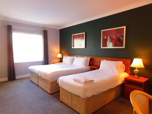 Habitación de hotel con dos camas, una ventana con cortinas, una mesita de noche con lámpara y dos cuadros colgados en la pared.