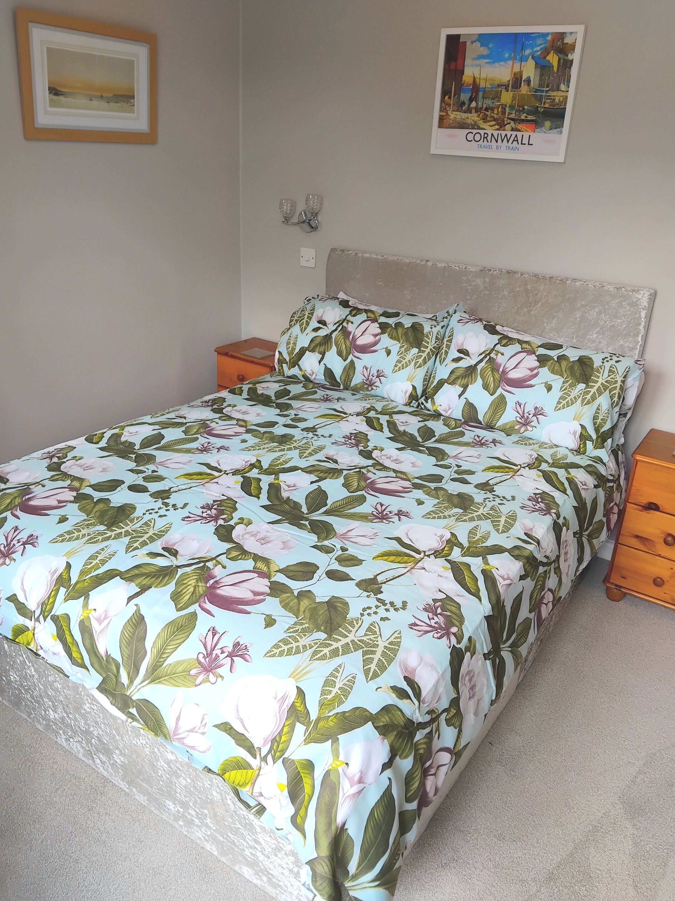 Ein Bett mit blumigem Bettzeug, einem Kopfteil, einem Nachttisch, einer Wandlampe und einem gerahmten Bild von Cornwall.