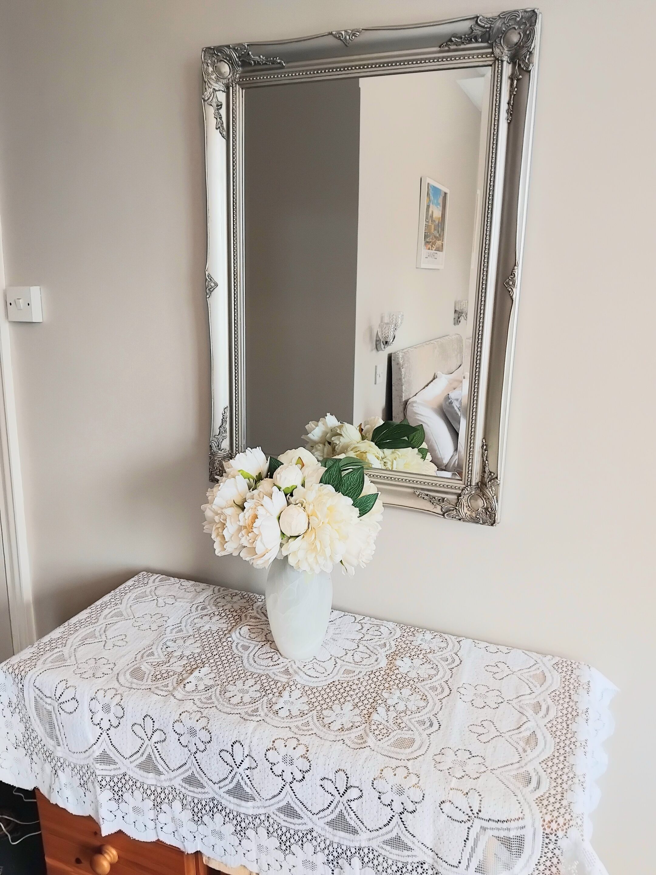 A mirror with an ornate frame, a lace tablecloth, and a vase with flowers on a table.