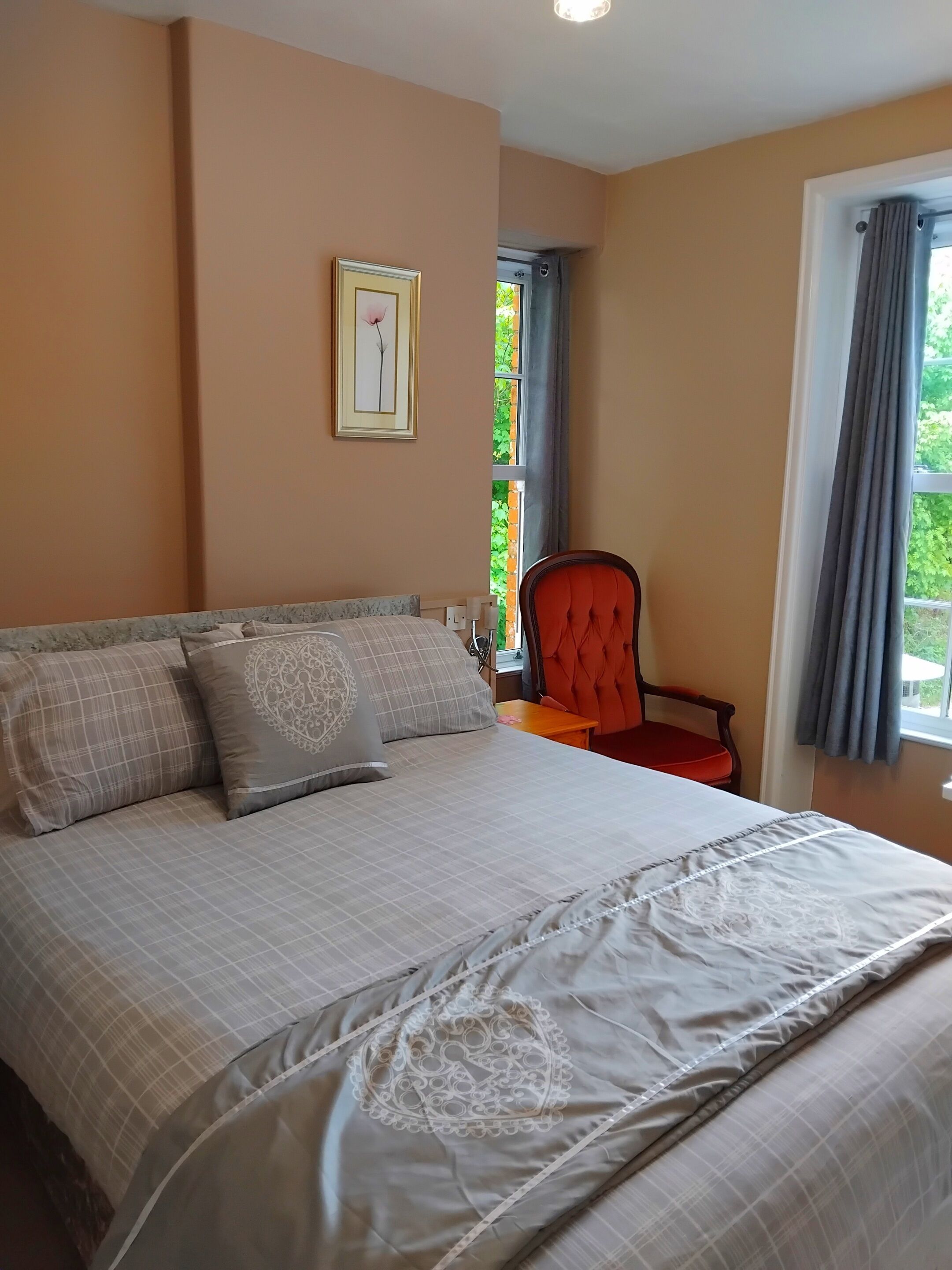 Ein Schlafzimmer mit Bett, Stuhl, Fenster mit Vorhängen und einem Bild an der Wand.