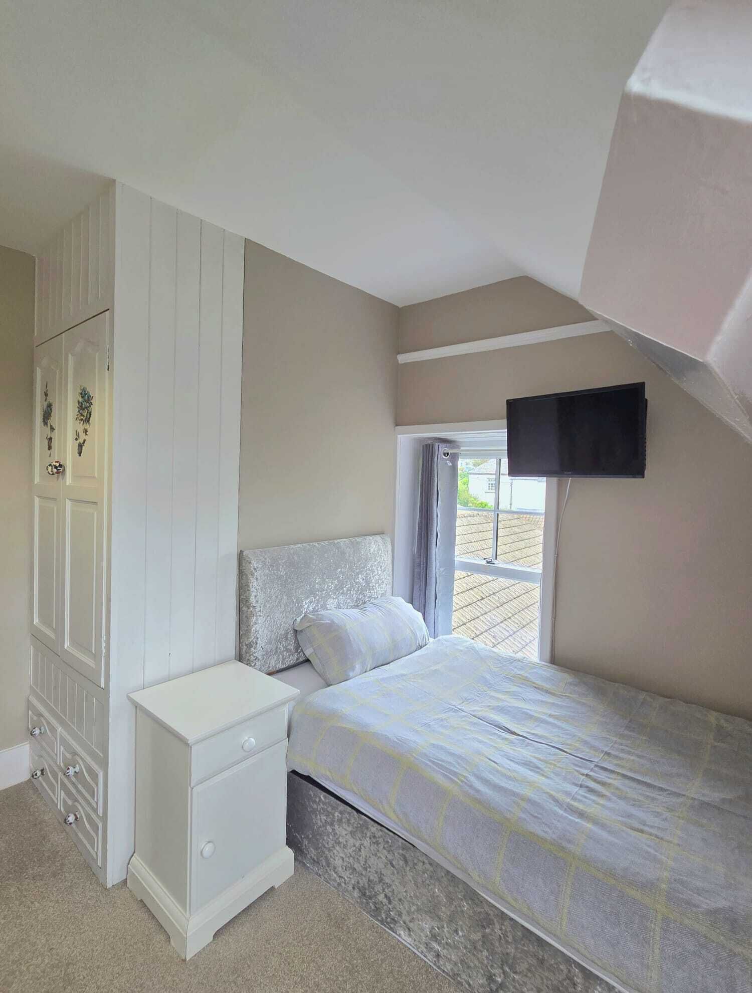 A bedroom with a bed, a nightstand, a wardrobe, a window with curtains, and a television mounted on the wall.