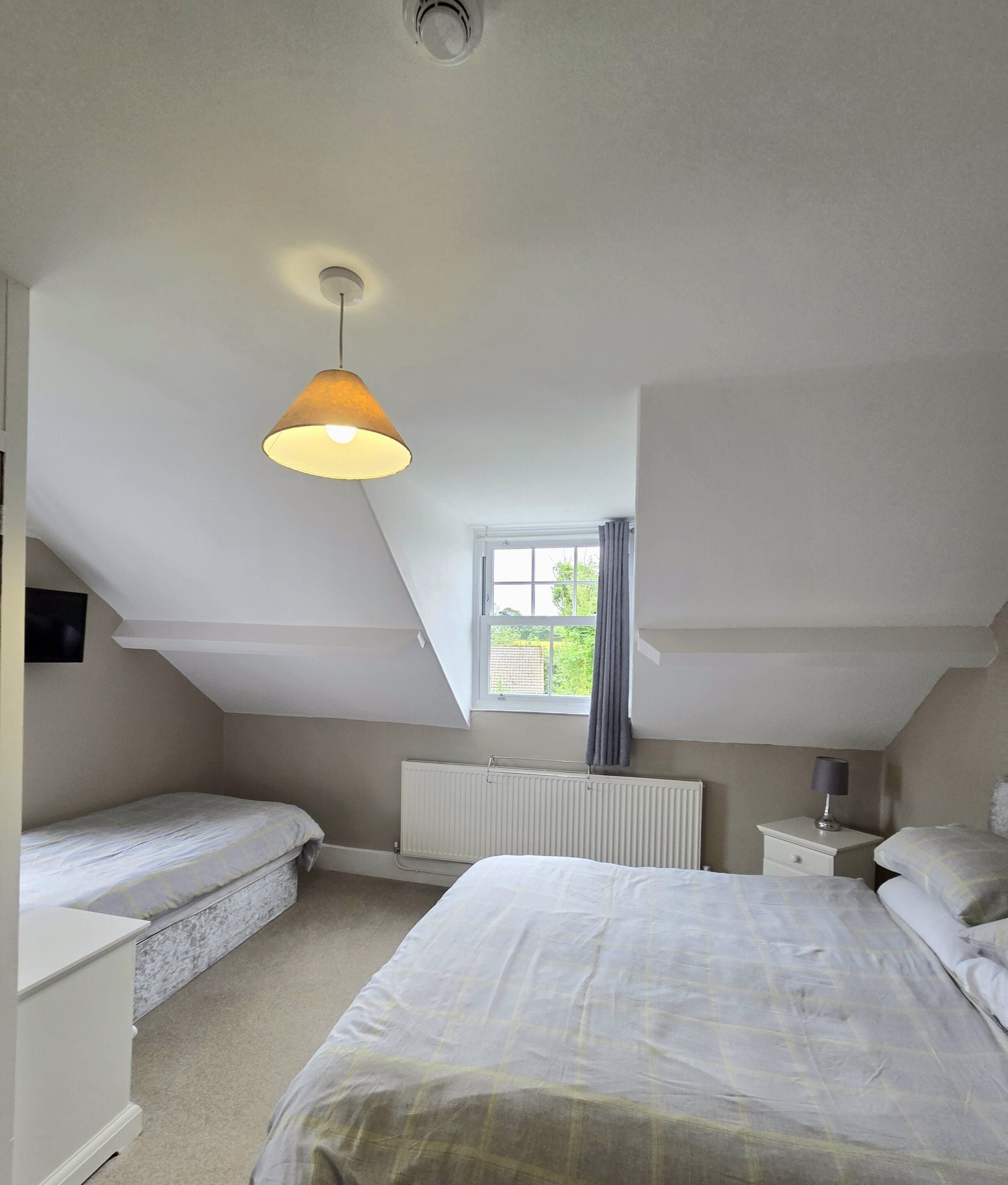 A bedroom with a sloped ceiling, a window with curtains, a radiator, a bed, a bedside table, and a hanging lamp.