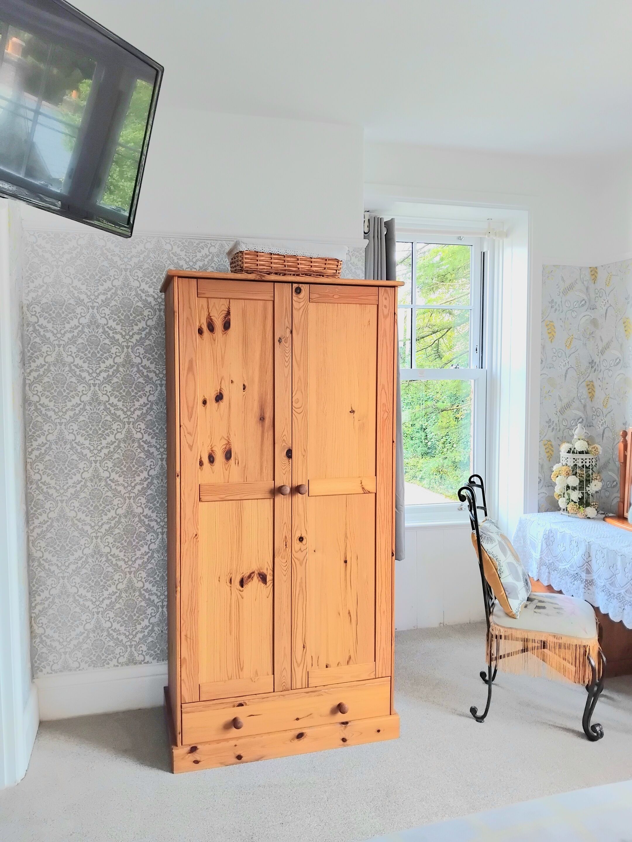 A wooden wardrobe with a basket on top, a chair with a cushion, and a window with a view of greenery.