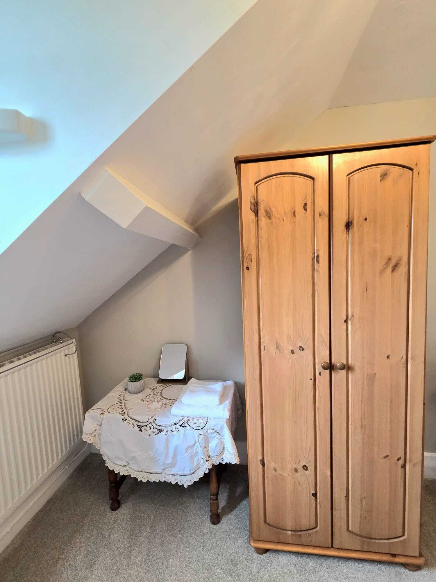 A bedroom with a sloped ceiling, a wooden wardrobe, a bedside table with a lamp, and a bed with a patterned bedspread.