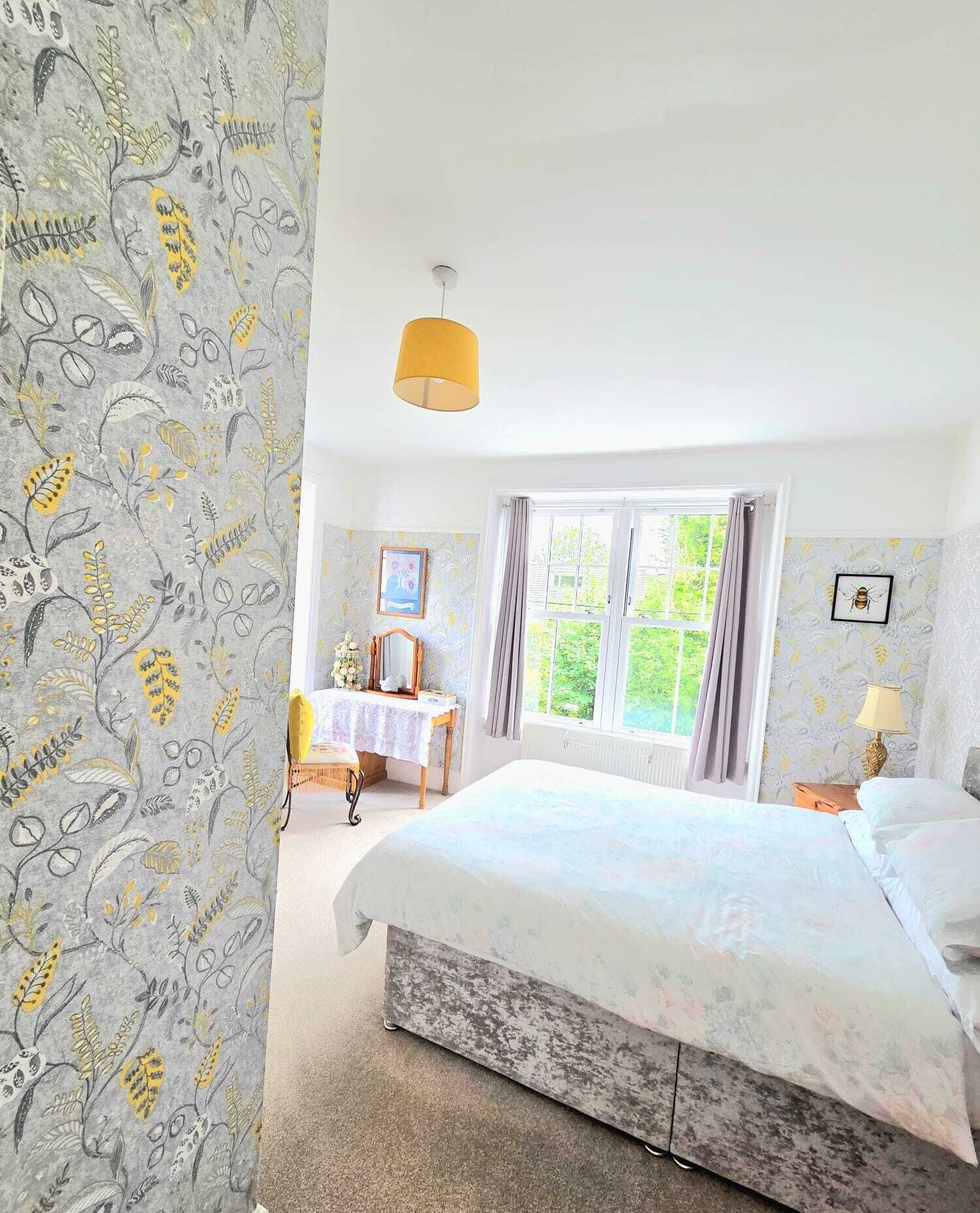 A bedroom with a bed, a bedside table, a window with curtains, a hanging lamp, and a floral wallpaper pattern.