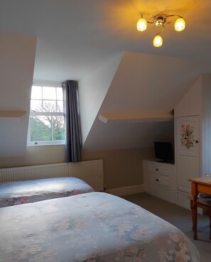 A bedroom with two beds, a radiator, a window with curtains, a desk with a chair, and a television on a dresser. - The Countryman Hotel (Camelford)