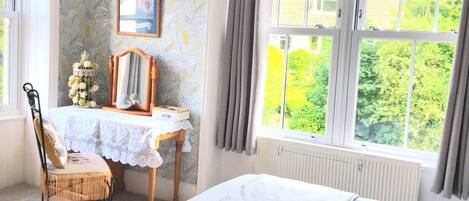 A bedroom with a bed, a desk, a chair, a radiator, and a window with a view of greenery.