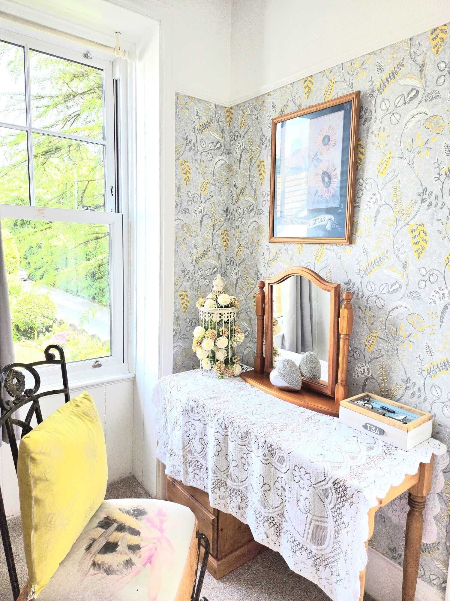 A room with a window, a desk with a lace tablecloth, a mirror, a framed picture, and a chair with a yellow cushion.