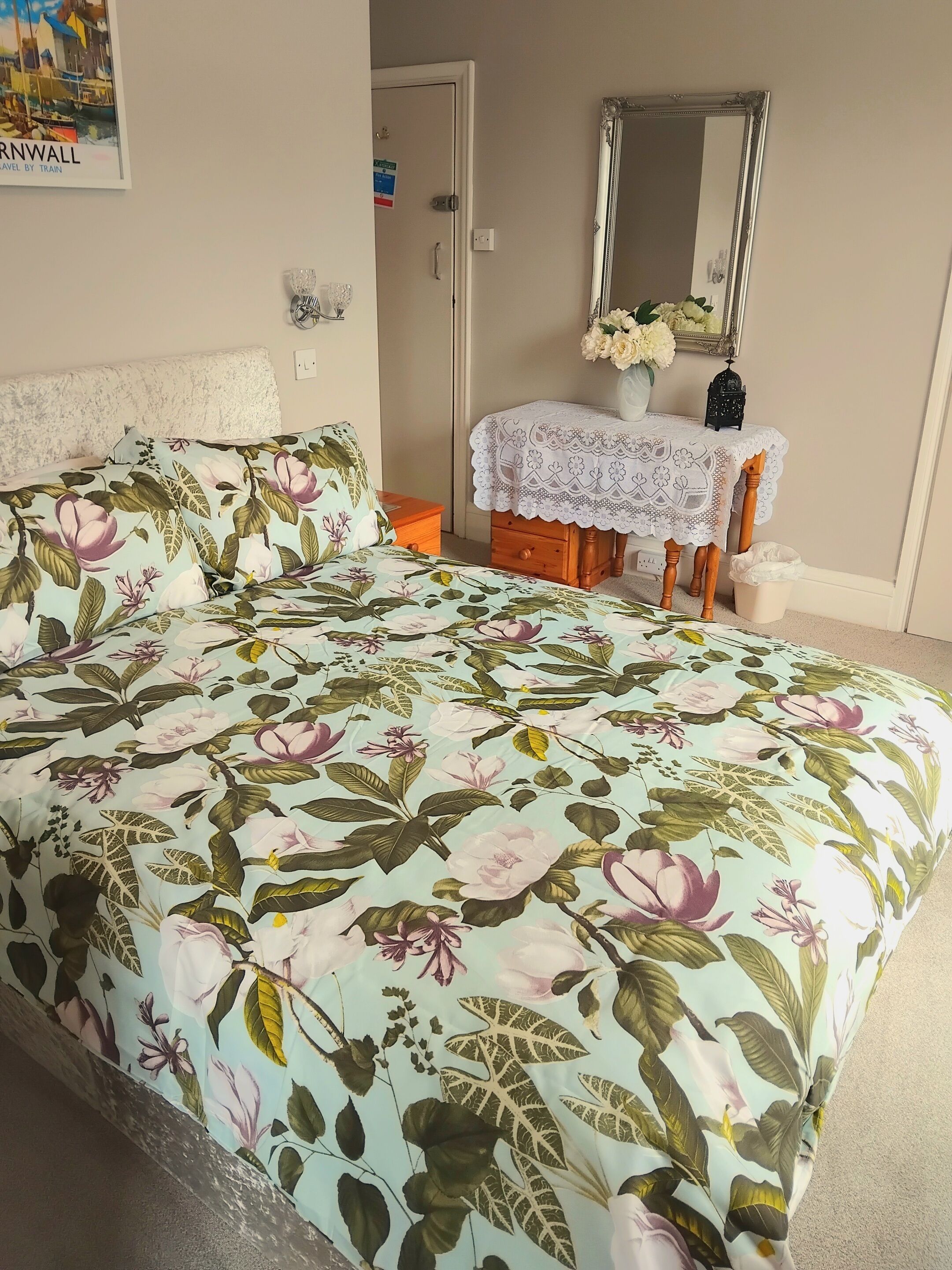 A bedroom with a floral bedspread, a bedside table with a vase, and a mirror on the wall.