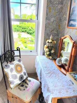 A room with a view of a garden, a chair with a patterned cushion, a table with a lace tablecloth, and a decorative birdcage with flowers. - The Countryman Hotel (Camelford)
