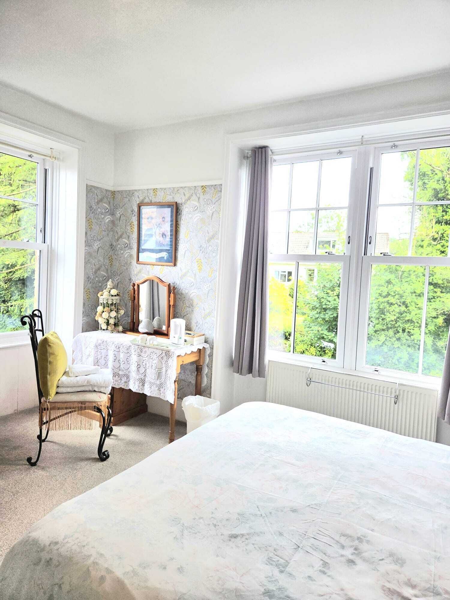 Un dormitorio con cama, escritorio, silla, radiador y ventanas amplias con vistas a zonas verdes.