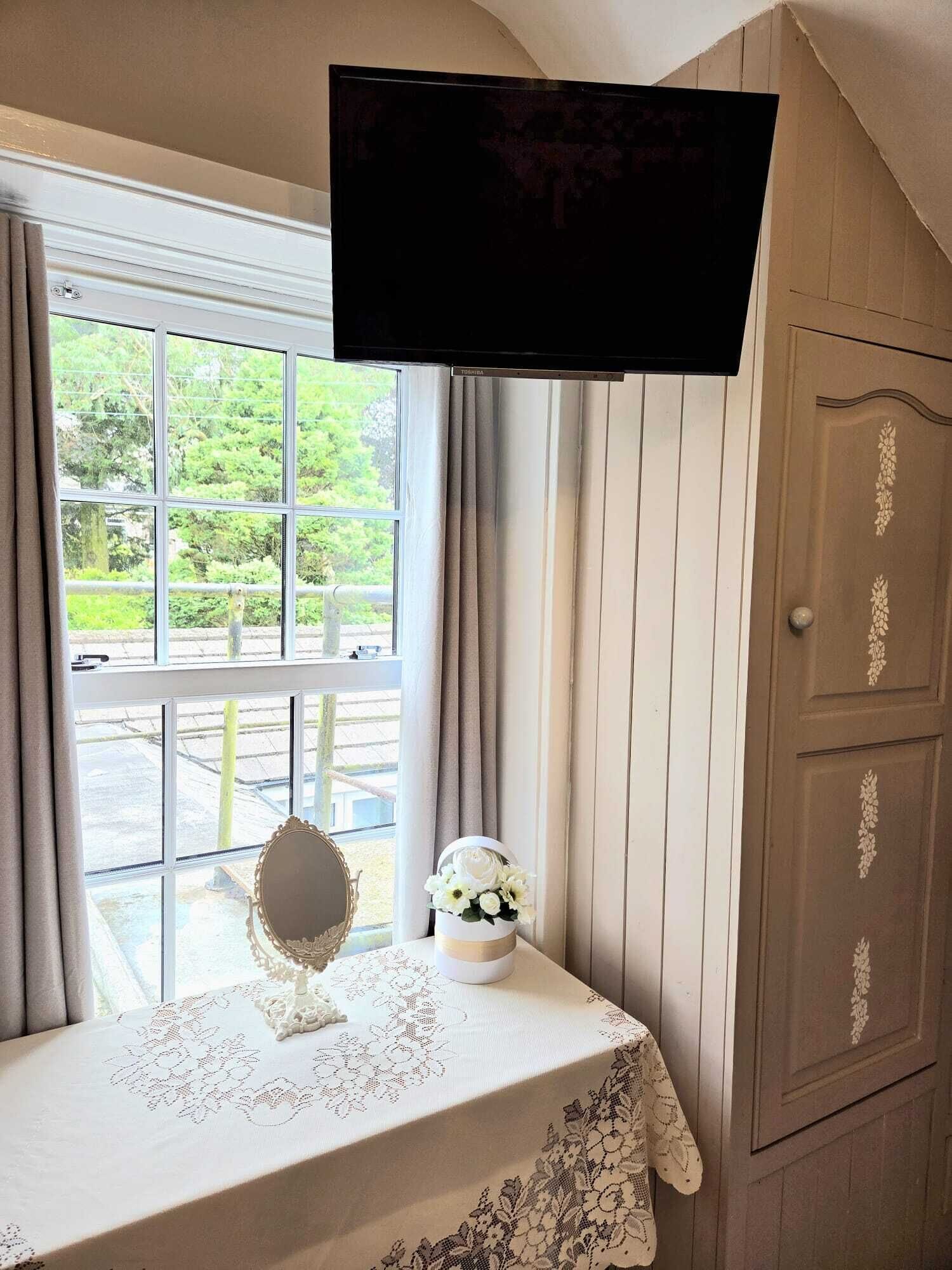 A room with a television mounted on the wall, a window with curtains, a table with a lace tablecloth, a mirror, and a decorative vase with flowers.