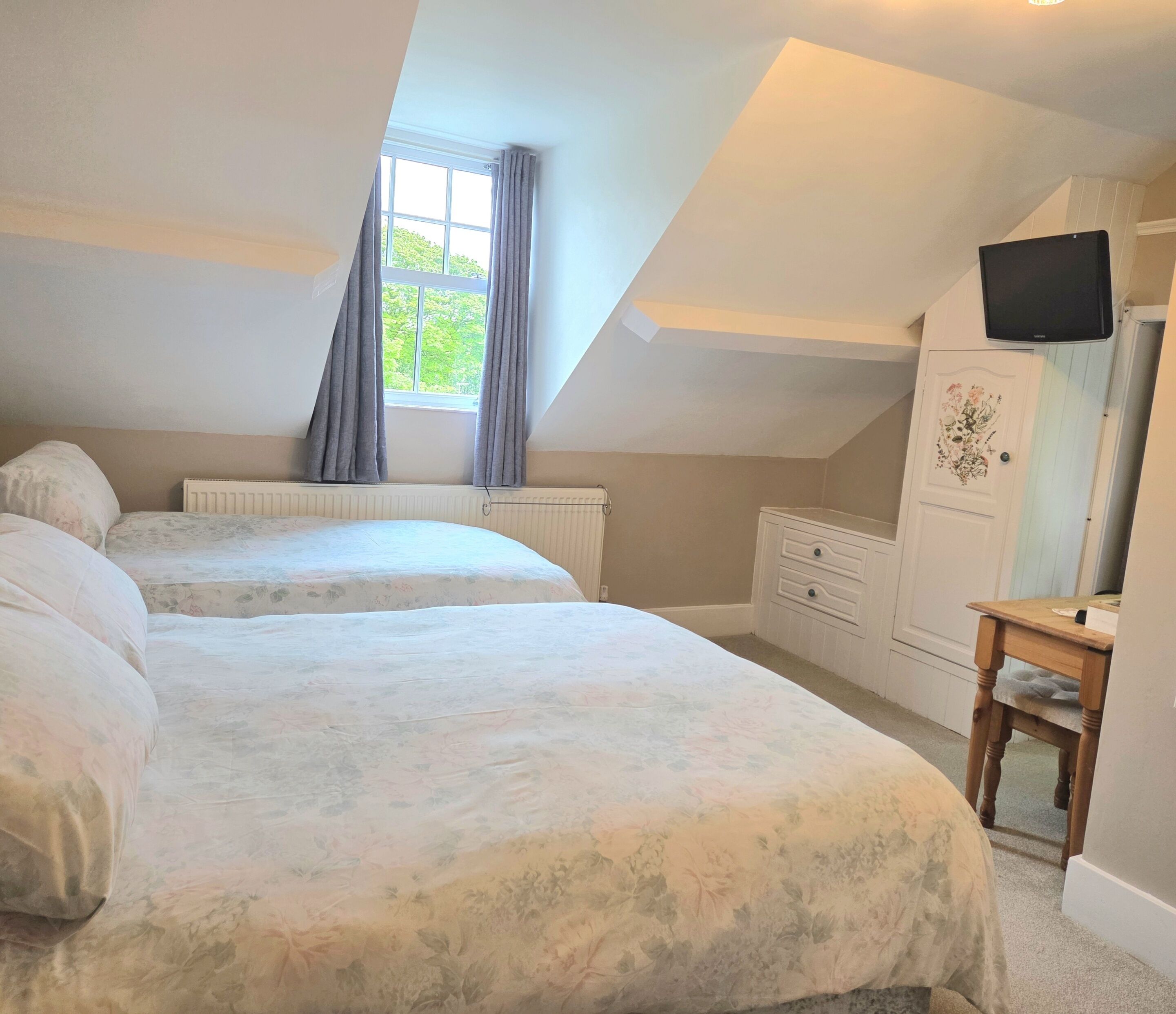 A bedroom with two beds, a window with curtains, a radiator, a wooden dresser, and a television on the wall.