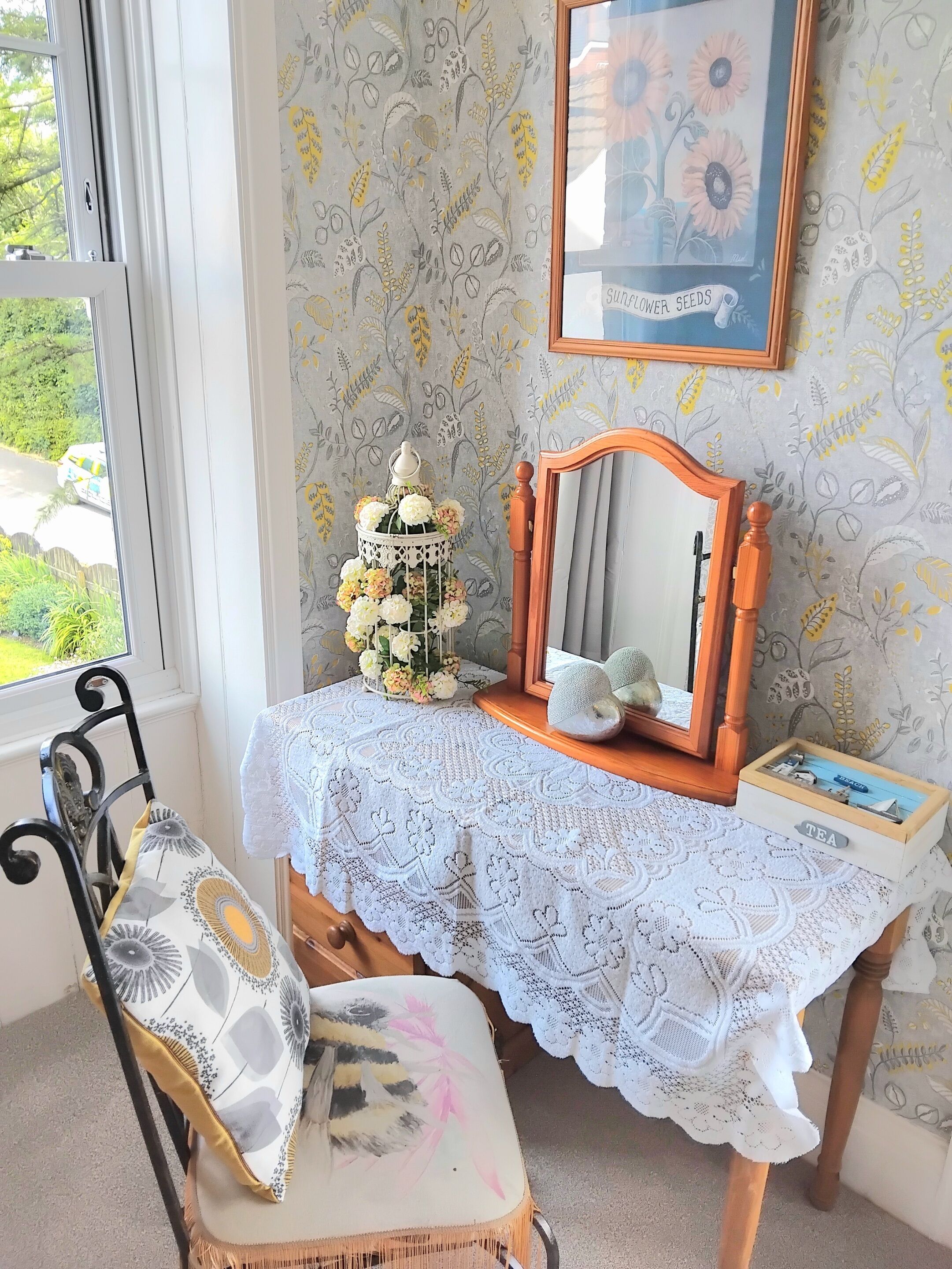 Une pièce comprenant une chaise en bois, une petite table recouverte d’une nappe en dentelle, une cage à oiseaux décorative, un tableau encadré et une fenêtre donnant sur un paysage verdoyant.