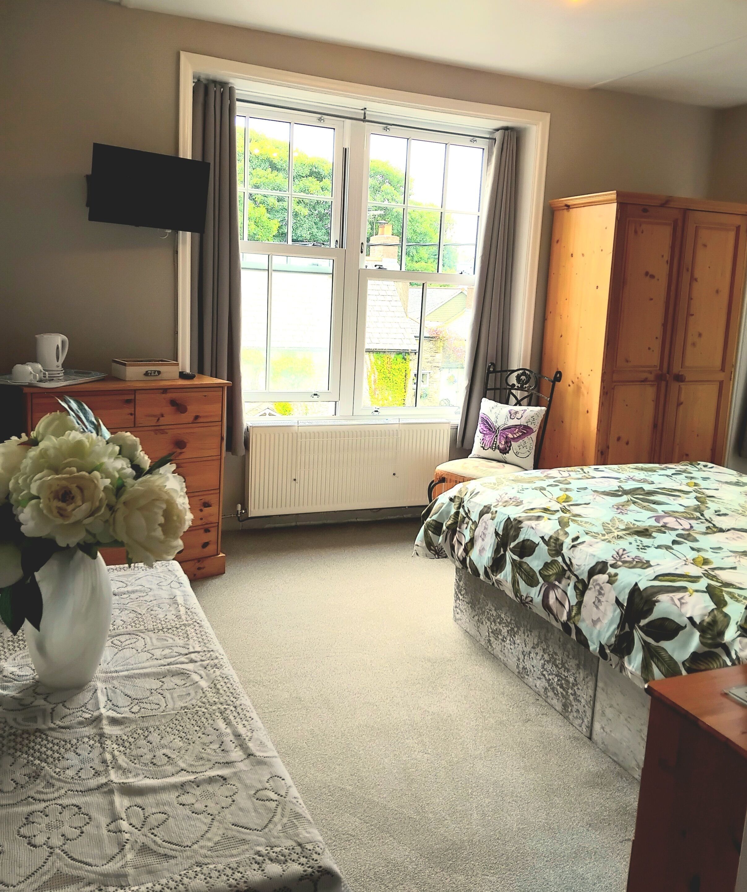A bedroom with a bed, a dresser, a television, a window with curtains, and a vase with flowers.