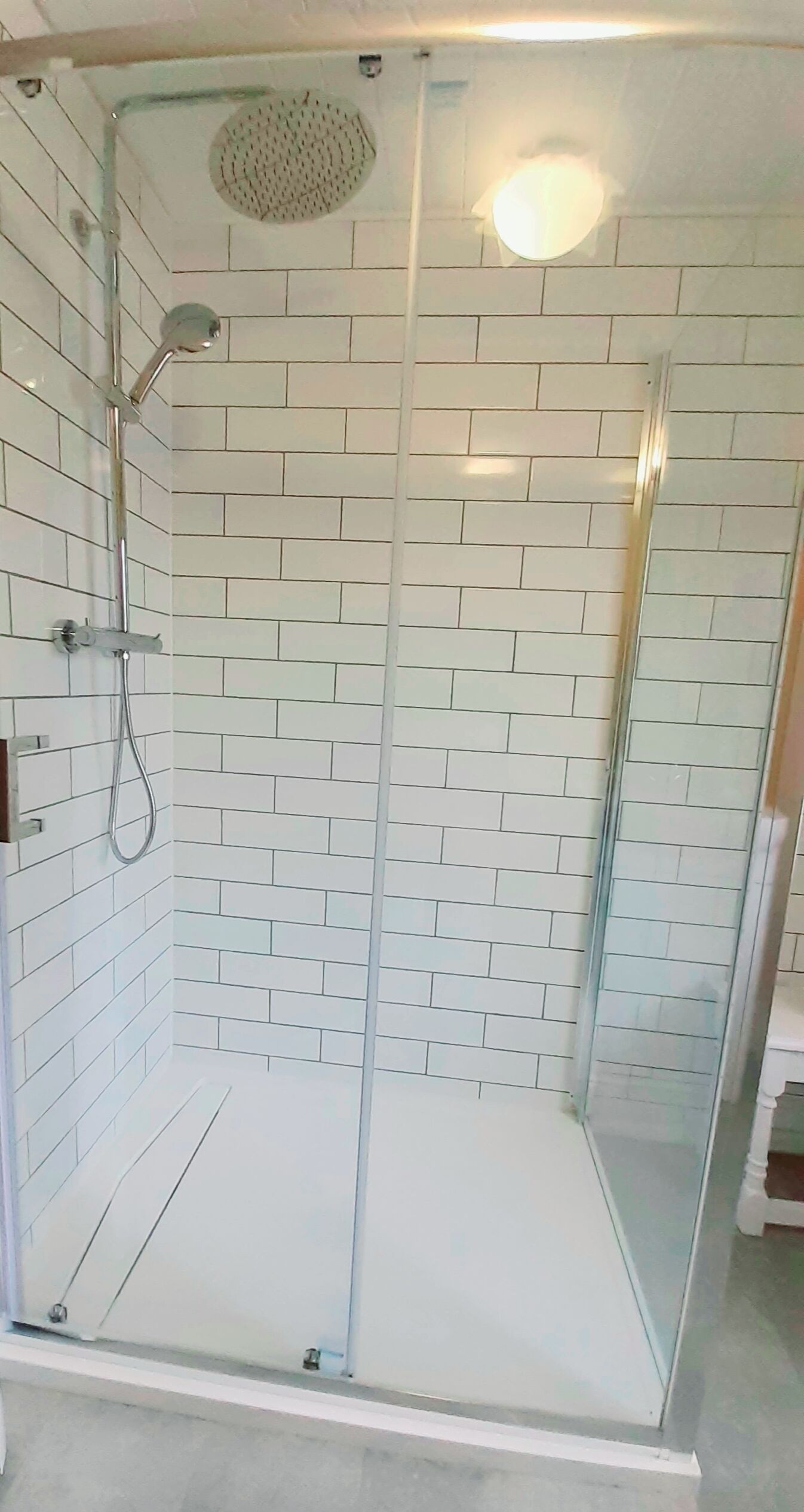 Une douche fermée par des parois de verre, avec des murs recouverts de carreaux blancs et un luminaire au plafond.