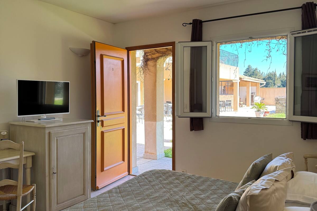 A bedroom with a bed, a TV on a cabinet, a desk with a chair, and a window with curtains.