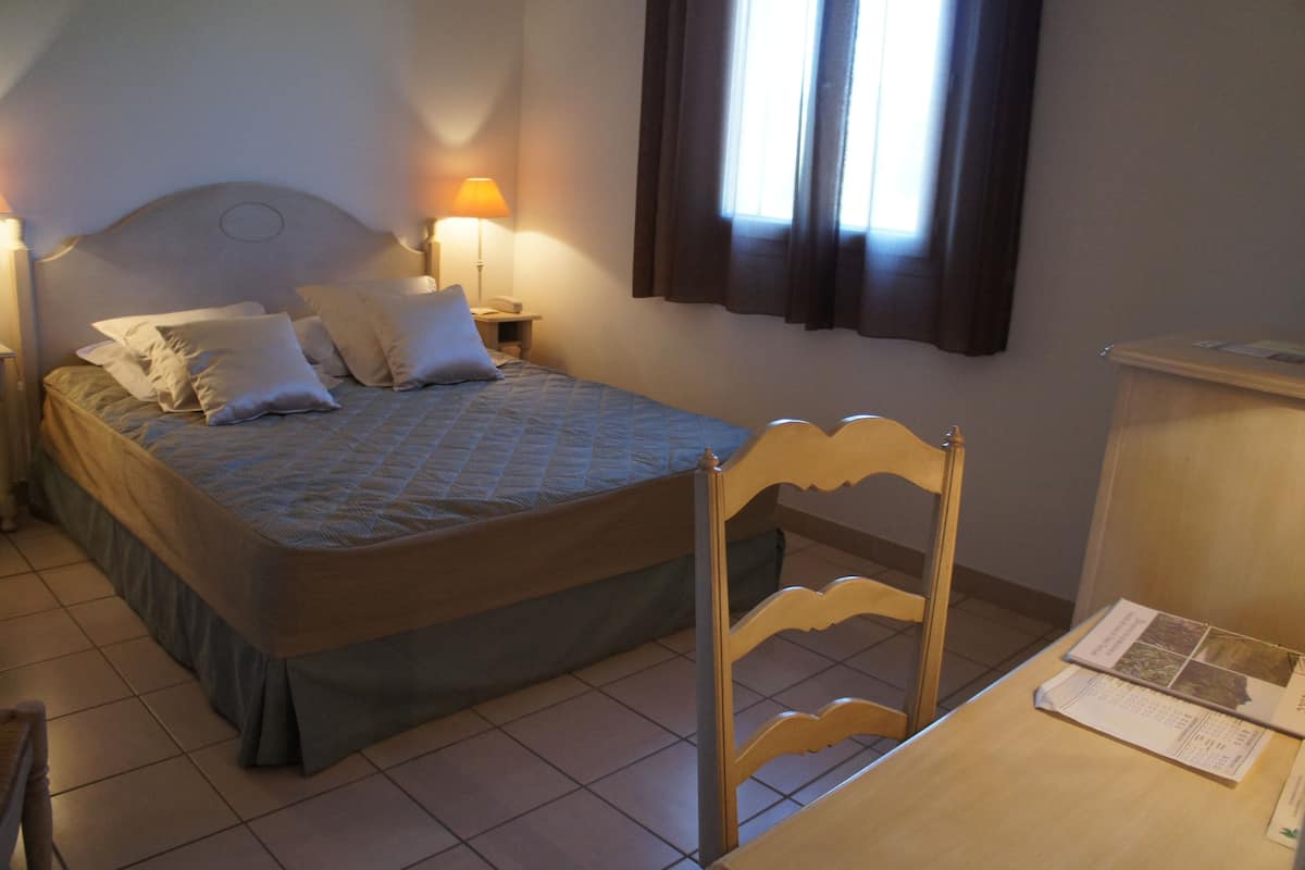 A neatly made bed with pillows, a bedside lamp, and a wooden chair at a desk with a brochure.