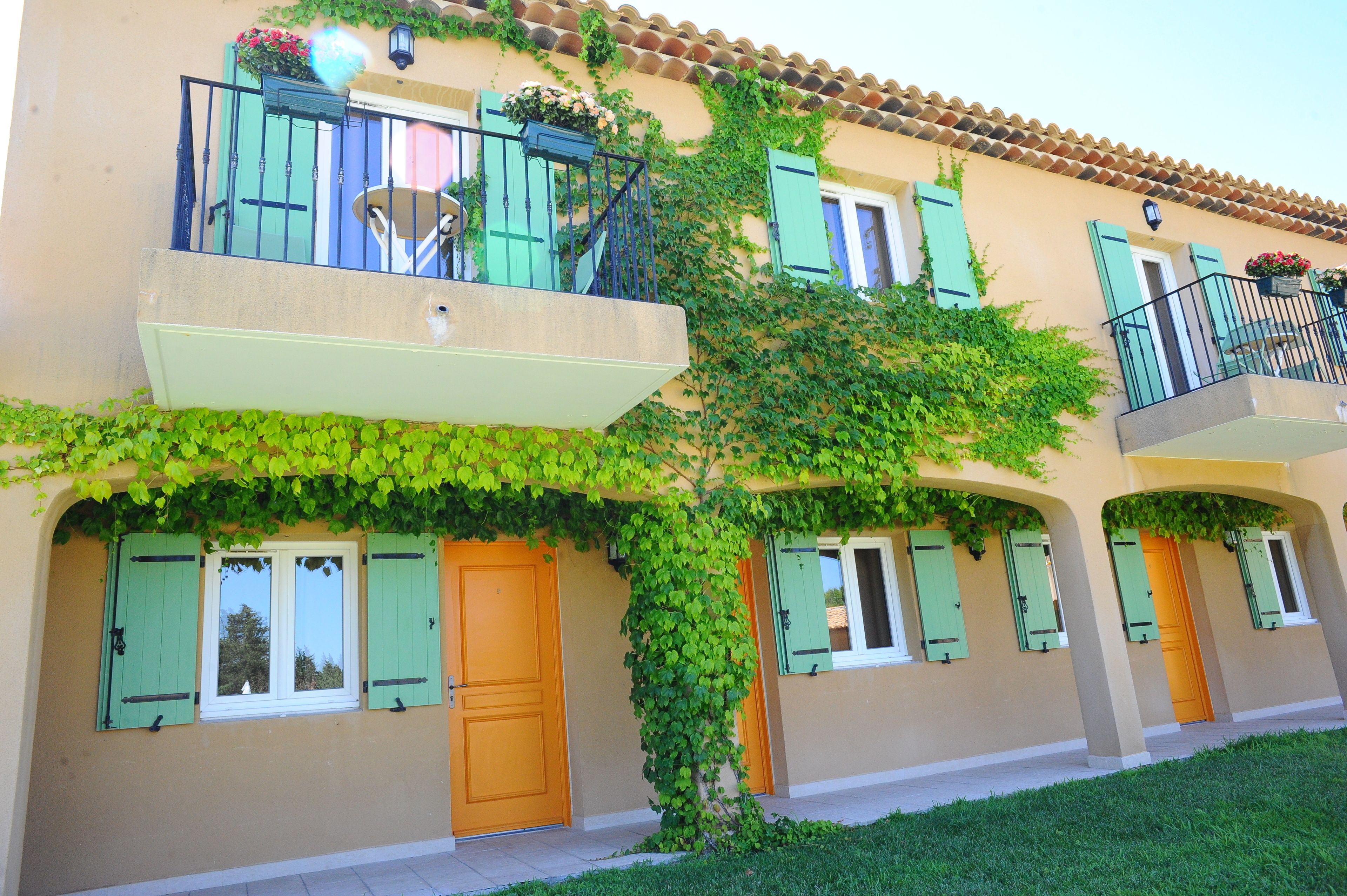 A two-story building with green shutters, a balcony with a chair, and a tree with green leaves growing on the facade.