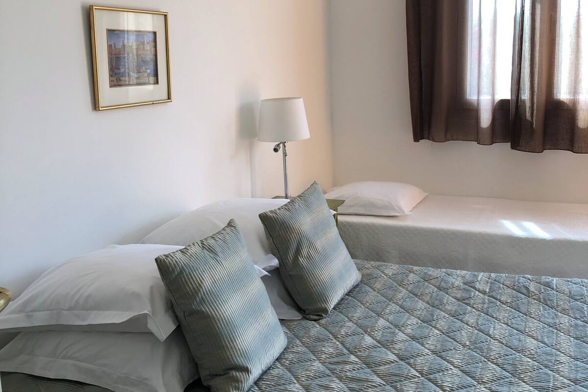 A bedroom with two beds, a framed picture on the wall, a bedside lamp, and a window with curtains.