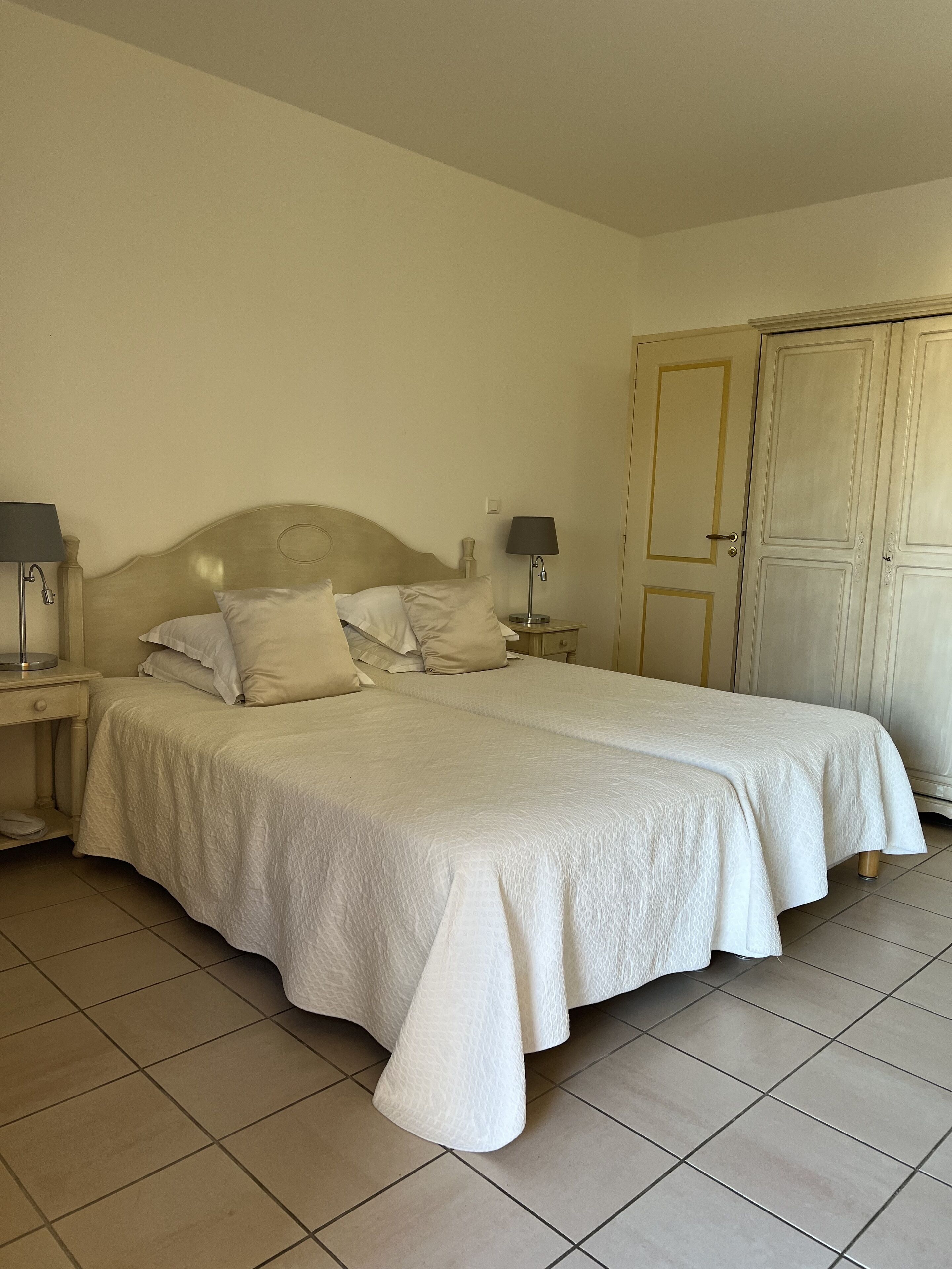 A double bed with a wooden headboard, two bedside tables with lamps, a wardrobe, and tiled flooring.