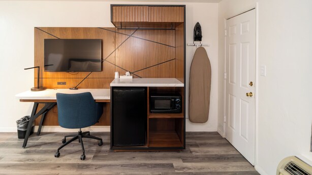 Une pièce de taille modeste, comprenant un bureau en bois, une télévision, un four à micro-ondes, un petit réfrigérateur, une chaise de bureau bleue et une porte blanche.