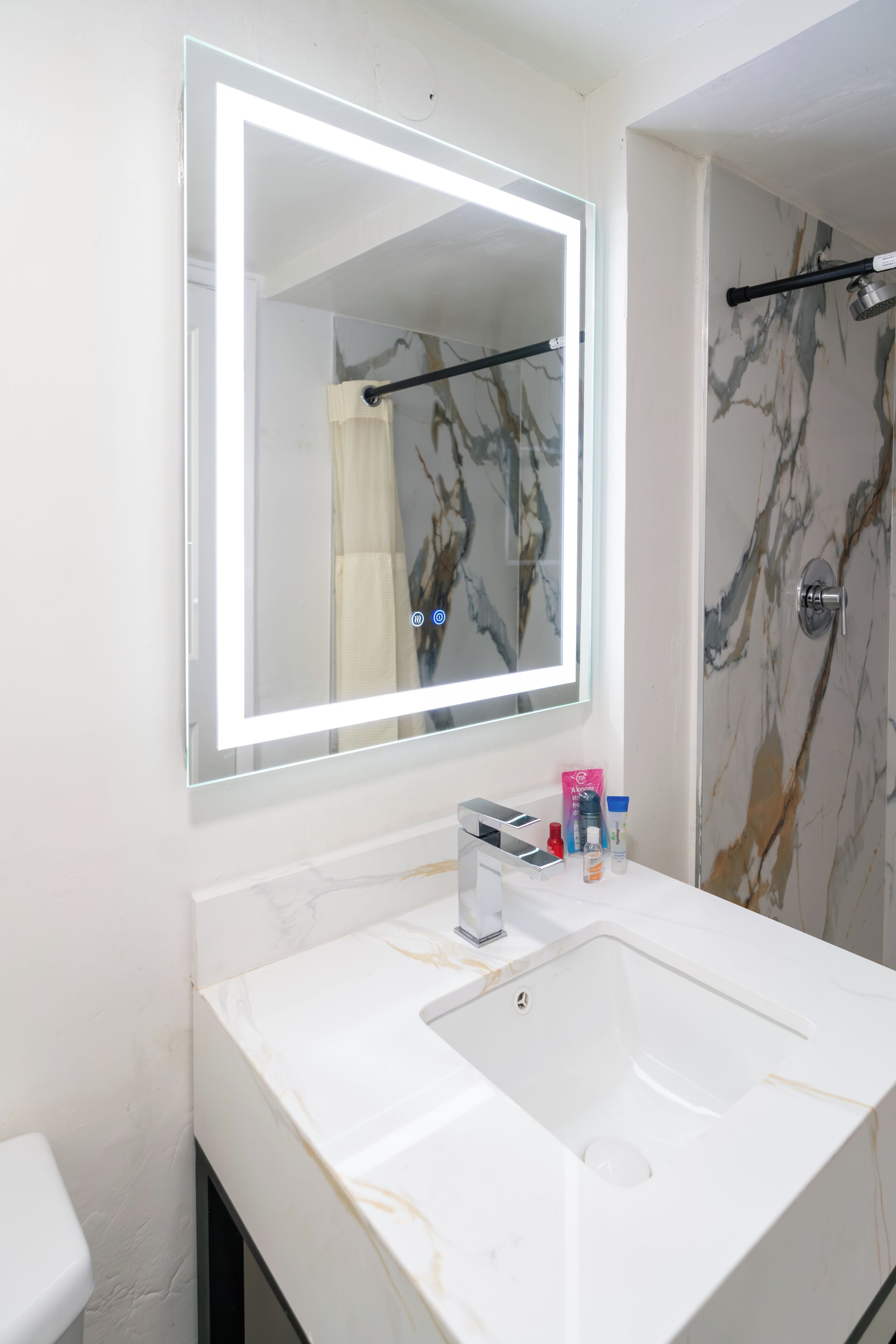 A bathroom with a white sink, a large mirror with a light, and a shower door with a marble pattern.